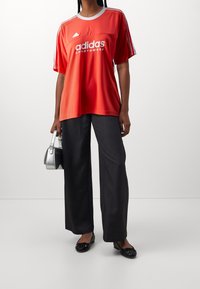 Červené sportovní tričko Adidas s bílým logem a proužkovými akcenty; kombinováno s tmavými širokými kalhotami a černými plochými botami. Součástí je stříbrná kabelka.