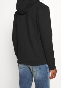 Felpa nera con cappuccio realizzata in un tessuto morbido e pieno di texture, maniche lunghe e tasca a marsupio, abbinata a jeans in denim azzurro chiaro.