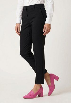 Frau trägt schwarze maßgeschneiderte Hose, weißes Hemd und leuchtend pinke Loafers mit Blockabsatz, steht mit gekreuzten Beinen.