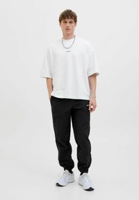 T-shirt blanc oversized à manches courtes avec du texte, associé à un pantalon de jogging noir avec des poignets élastiques et des poches, et des chaussures de sport blanches.