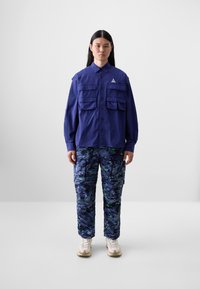 Lilla langermet skjorte med knappelukking og fire frontlommer. Blå kamuflasje-bukser med grønne detaljer og cargo-lommer. Hvite joggesko.