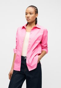 Γυναίκα με πλεξούδες, φορώντας ένα ροζ πουκάμισο με ρίγες κουμπωμένο πάνω από μπεζ τοπ και σκούρα μπλε τζιν, στέκεται με το ένα χέρι στην τσέπη.