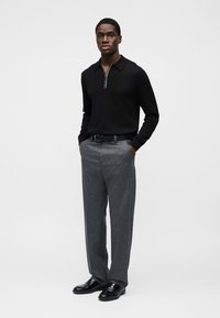 Μαύρο πλεκτό πόλο μπλουζάκι με φερμουάρ στο λαιμό. Γκρι ριγωτές παντελόνες και μαύρα γυαλιστερά παπούτσια. Το μοντέλο στέκεται με τα χέρια στις τσέπες σε λευκό φόντο.