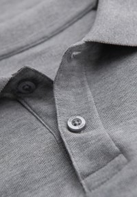 Närbild av ljusgrått texturerat tyg med en knapp fäst på en knappslå, som visar detaljer av söm och vävning.