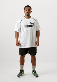 White, short-sleeve jersey with black Puma logo, paired with black patterned shorts, gray socks, and green accent sneakers.