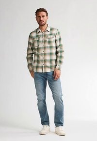 Chemise en flanelle à carreaux verts et crème avec deux poches poitrine, fermeture à boutons et manches longues, associée à un jean bleu clair et des baskets blanches.