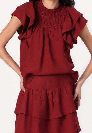 Woman wearing a sleeveless dark red top with ruffled shoulders and a matching tiered skirt, hands resting on hips.