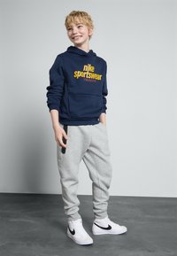 Tumši zila kapuce ar dzelteno "Nike Sportswear" logotipu; pelēkas treniņbikses ar sānu kabatām; baltas snickers ar melnām akcentiem. Gluda auduma tekstūra.