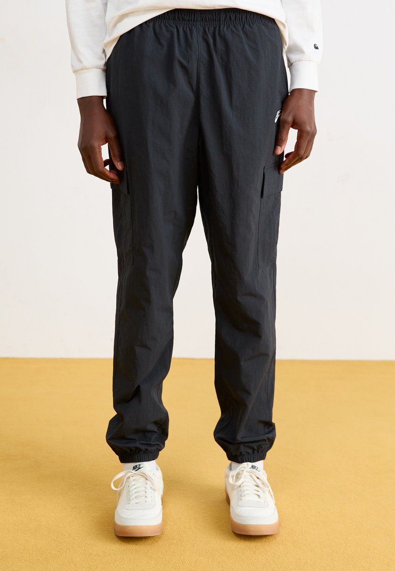 Pantalons cargo noirs avec une taille élastique, des poches latérales et des poignets fuselés. Fabriqués en tissu léger. Portés avec des baskets blanches.