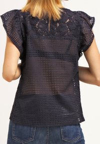 Haut bleu marine avec détails en dentelle sur les épaules et dos en mesh transparent à motif quadrillé. Manches courtes volantées et coupe décontractée.