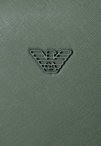 Strukturēts zaļš materiāls ar subtilu spīdumu, centrā ir tumšs logotipa dizains ar stilizētām spārniem.