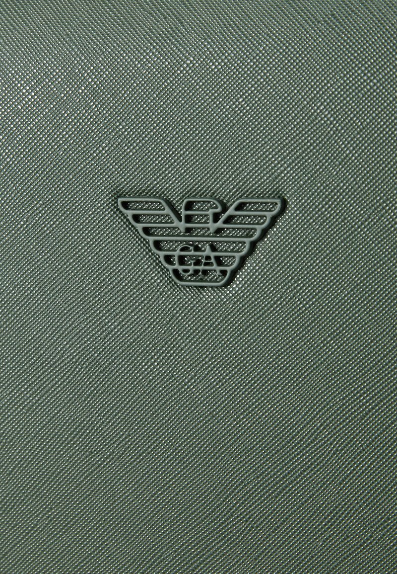 Material verde texturizado con un brillo sutil, que presenta un diseño de logo oscuro de alas estilizadas en el centro.