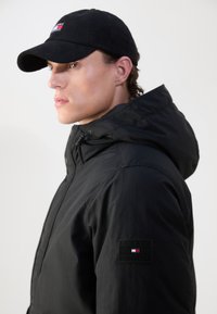 Černá kapucňová bunda s hladkou texturou a nášivkou s logem Tommy Hilfiger na rukávu. Černá kordová čepice dotváří celkový vzhled.