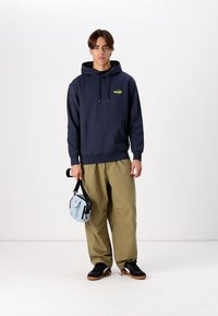 Navy hoodie with front pocket, green logo, khaki pants, black shoes, and a light blue crossbody bag with black strap.