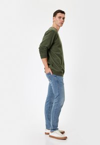 Grøn langærmet skjorte, denimjeans, hvide sneakers med brune såler. Modellen står på siden, med hænderne i lommerne, neutral baggrund.