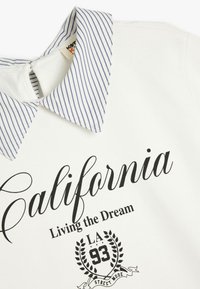 Sudadera blanca con un cuello a rayas en azul y blanco, que presenta el texto "California Living the Dream" y un acento gráfico en negro.