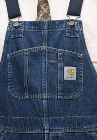 Μπλε τζιν φορμάκι Carhartt με μπροστινή τσέπη, μεταλλικά κούμπωμα, φορεμένο πάνω από λευκό πουκάμισο και καφέ γραβάτα με σχέδια.