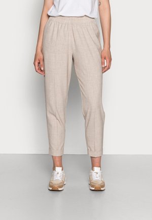 Mujer con pantalones beige hasta el tobillo con puños remangados, zapatillas blancas y una camiseta blanca metida por dentro, de pie contra un fondo liso.