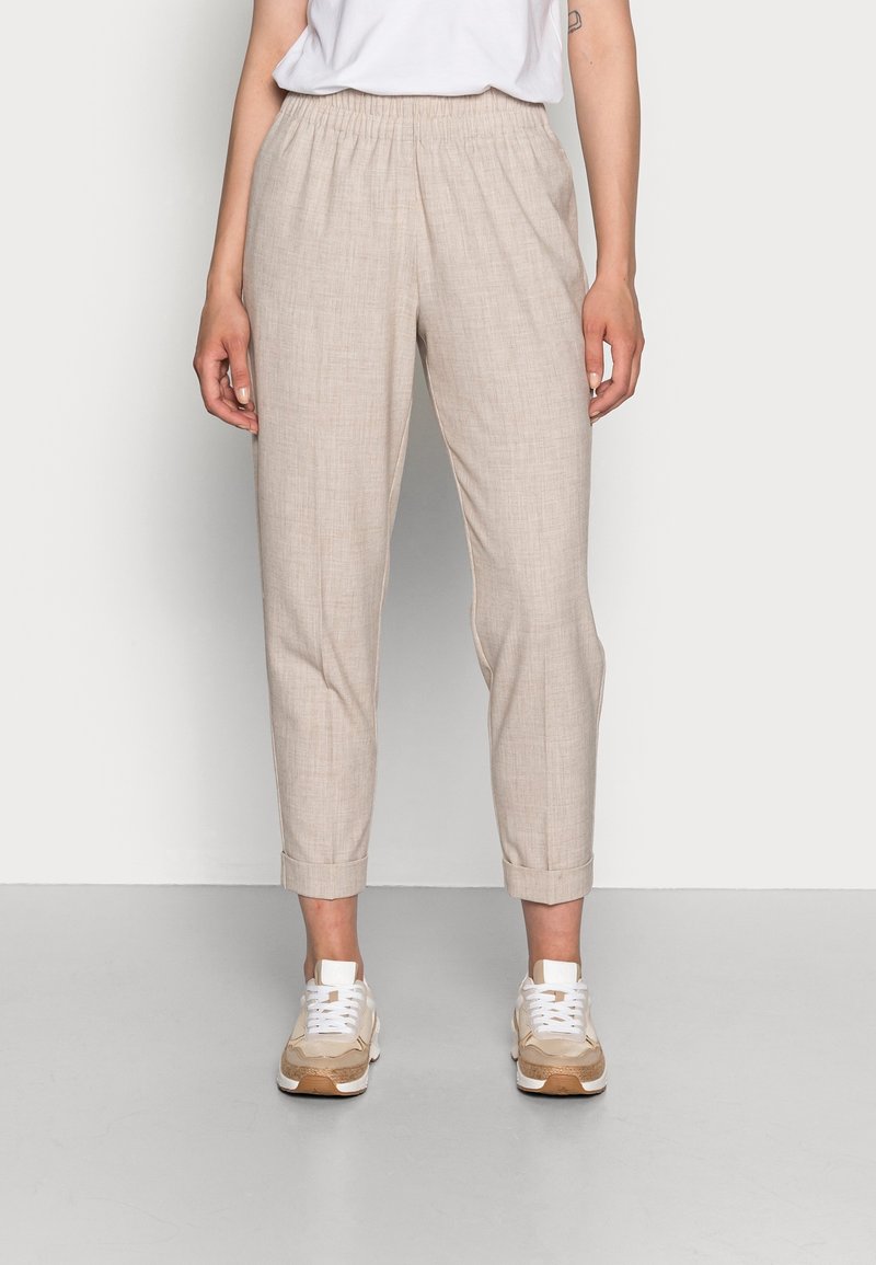 Femme portant un pantalon beige longueur cheville avec des revers, des baskets blanches et un t-shirt blanc rentré, debout devant un fond uni.
