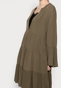 Olivgrön, lager-på-lager klänning i lätt material, med V-ringning och trumpetformade långa ärmar. Mjuk textur med en svävande silhuett.