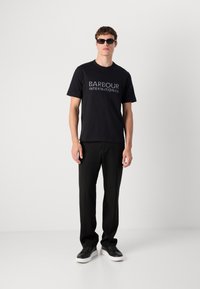 T-shirt preta com impressão "BARBOUR INTERNATIONAL", combinada com calças pretas e ténis pretos com solas brancas, usados por uma pessoa.