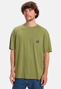Olive green cotton t-shirt with short sleeves, round neckline, and chest pocket featuring a black logo patch. Casual fit and lightweight texture.