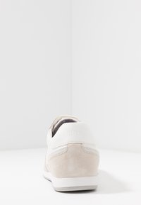 Bota s bílým koženým svrškem a béžovými semišovými akcenty. Obsahuje texturovaný design, polstrovaný límec a nenápadné logo na patě.