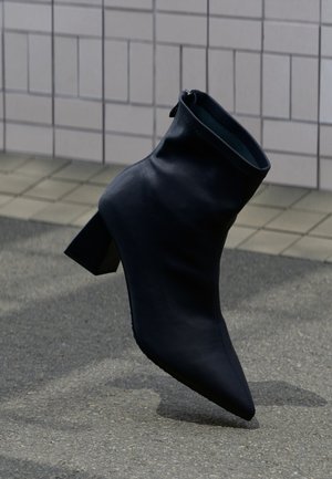 Bottine noire en cuir avec talon bloc et bout pointu, posée sur une surface carrelée et asphaltée.