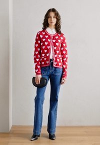 Punainen neulottu neuletakki, jossa on valkoinen sydänkuvio, napitus ja ribbireunus. Yhdistetty sinisiin leveälahkeisiin farkkuihin ja mustaan käsilaukkuun, neutraalin taustan edessä.