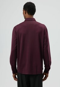 Donker bordeauxrode shirt met lange mouwen, klassieke kraag, gladde textuur en knoopmanchetten, gezien van achteren tegen een neutrale achtergrond.