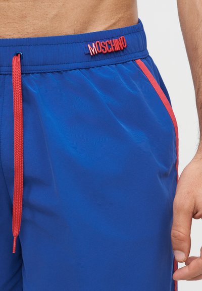Nærbilde av blå badeshorts med rød snor og rød MOSCHINO-logo på midjebåndet, båret av en person med synlig hånd.