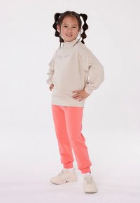 Šviesiai smėlio spalvos džemperis su aukštu apykakliu ir išsiuvinėtu užrašu, derinamas su ryškiai rožinėmis laisvalaikio kelnėmis ir šviesiais sportbačiais. Lygi medžiaga ir laisvas siluetas.