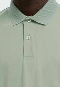 Lysegrønn poloskjorte med en teksturert mesh-krage, som har to knapper og et glatt stoff. Enkel og klassisk design, nærbilde.