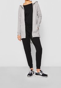 Persona con un hoodie grigio con zip, pantaloni jogger neri e sneakers New Balance bianco e nero, in piedi contro uno sfondo bianco semplice.