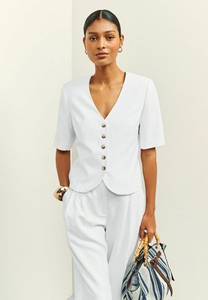 Kobieta z zaczesanymi do tyłu włosami nosi białą zapinaną na guziki bluzkę i spodnie, nosząc niebiesko-białą paskowaną torebkę z drewnianymi uchwytami.