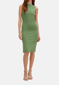 Femme portant une robe verte sans manches, ajustée, longueur genou, à rayures horizontales, et des sandales beige à talons hauts avec brides.