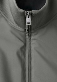 Zbliżenie na suwak szarej kurtki z żeberkowym kołnierzem i metalowym uchwytem z wygrawerowanym logo "MNG".