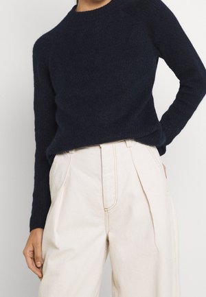 Person iført en mørk marineblå tekstureret langærmet sweater, stoppet ned i højttaljede, plisserede off-white bukser med synlige sømme.