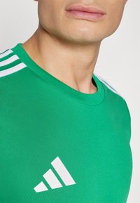 Green athletic shirt made of smooth fabric featuring a crew neckline, white three-stripe accents on the shoulders, and a white logo design.