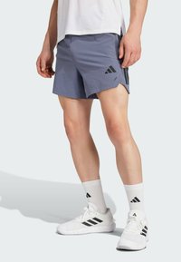 Pantaloni sportivi da uomo in tessuto azzurro, con uno spacco laterale, logo Adidas nero, abbinati a calzini sportivi bianchi e scarpe da ginnastica bianche.