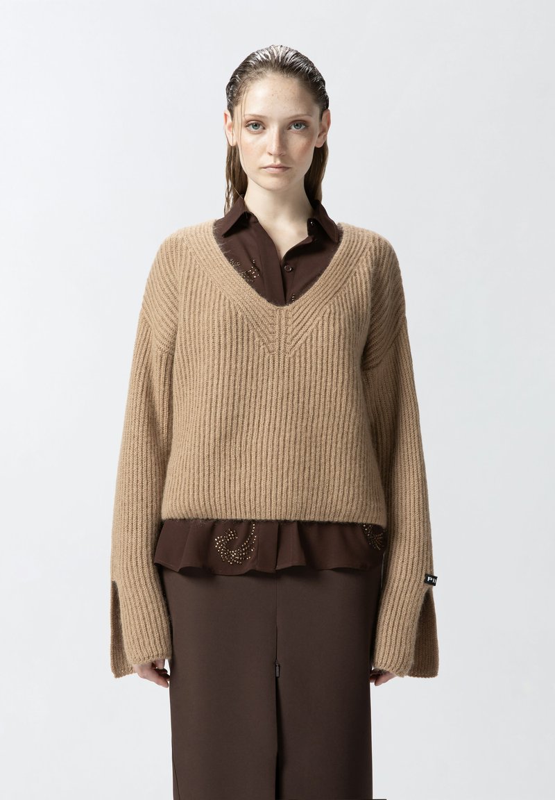Maglione a coste beige con scollo a V, dotato di maniche lunghe e spacchi laterali. Indossato sopra una camicia marrone scuro con volant e decorazioni.