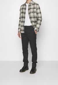 Uomo che indossa una camicia a quadri bianca e nera sopra una t-shirt bianca, pantaloni neri e stivali neri con lacci, in piedi su uno sfondo bianco semplice.