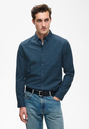Junger Mann mit kurzem braunem Haar, trägt ein dunkelblaues Hemd mit Knöpfen, das in helle blaue Jeans mit schwarzem Gürtel gesteckt ist, Hand in der Tasche.