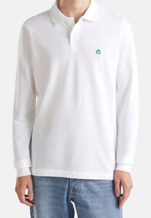 Hombre con camisa polo blanca de manga larga con un pequeño logo verde en el pecho y vaqueros azules, de pie frente a un fondo liso.
