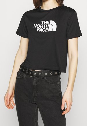 Schwarzes, kurzes T-Shirt mit weißem Logo "THE NORTH FACE." Es hat kurze Ärmel und eine glatte Textur, kombiniert mit dunkler Jeans und einem mit Nieten besetzten Gürtel.