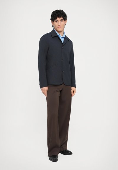 Tengerészkék gombos dzseki gallérral, barna nadrággal párosítva. A dzseki letisztult dizájnnal és két oldalzsebbel rendelkezik, puha anyagból készült.