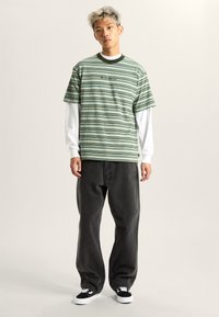 Stribet kortærmet grøn og hvid t-shirt lagdelt over en langærmet hvid skjorte, parret med mørke grå bukser og sorte sneakers.
