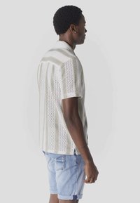 Homme portant une chemise tricotée beige et blanche à manches courtes à rayures, avec un short en denim bleu clair, debout de profil devant un fond uni.