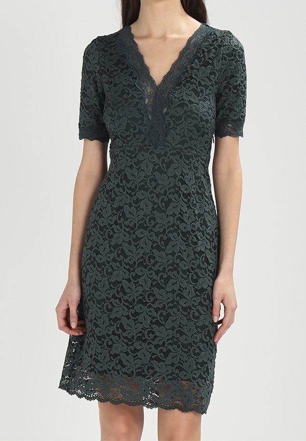 Femme portant une robe en dentelle vert foncé longueur genou avec des manches courtes et un décolleté en V festonné.
