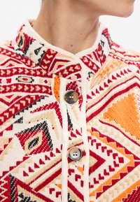 Mintás, szőtt dzseki geometriai mintákkal piros, narancs és krém színekben. Mandarin gallérral és két fémbottonnal rendelkezik.